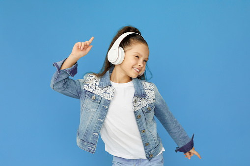 happy little child girl in white headphones and denim listening to music and dancing on blue background