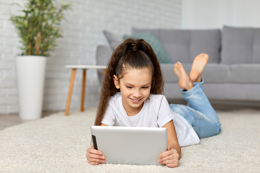 cute happy little child girl lying on the carpet and using digital tablet at home. kid enjoying tablet