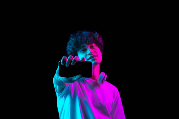 Young Caucasian man showing phone screen isolated over dark background in neon