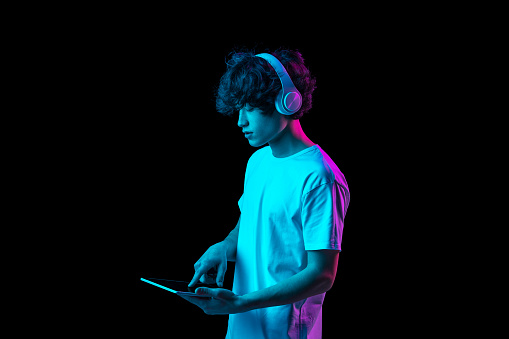 Digital world. Young man using tablet, device isolated over dark multicolored background in neon light. Future, gadgets, technology, education online concept. Human emotion, facial expressions. .
