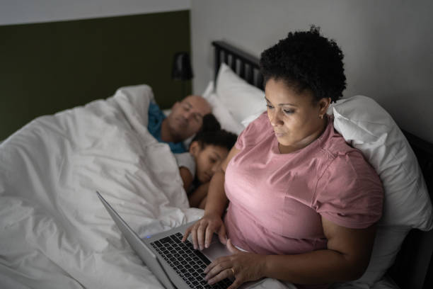 donna adulta media che usa laptop a letto mentre marito e figlia dormono - addiction internet computer working late foto e immagini stock