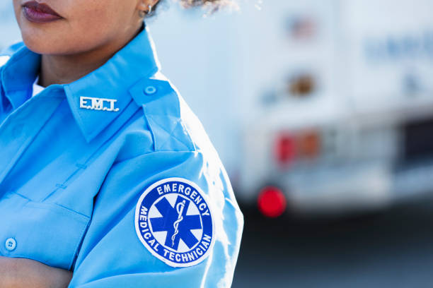 Female paramedic in front of ambulance Cropped view of a female paramedic standing with arms crossed in front of an ambulance. She is a mid adult woman in her 30s, mixed race Hispanic and Pacific Islander. The focus is on the generic EMT patch on her sleeve at the shoulder. emt stock pictures, royalty-free photos & images