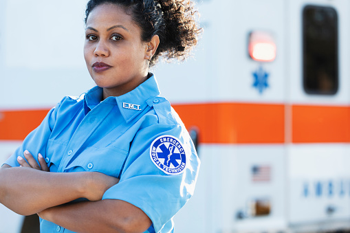 A female paramedic standing with arms crossed in front of an ambulance, looking at the camera. She is a mid adult woman in her 30s, mixed race Hispanic and Pacific Islander.