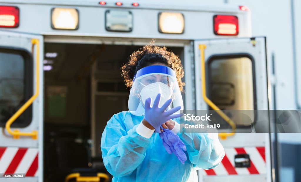 Paramedic putting on PPE during COVID-19 A paramedic standing in front of an ambulance, putting on PPE, or personal protective equipment, including a face mask, face shield, surgical gown and gloves. She is working during the COVID-19 pandemic, trying to protect herself from catching and spreading coronavirus. She is a mid adult woman in her 30s, mixed race Hispanic and Pacific Islander. Paramedic Stock Photo