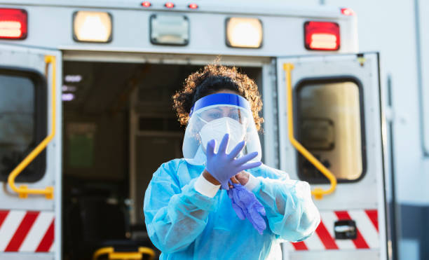 Paramedic putting on PPE during COVID-19 A paramedic standing in front of an ambulance, putting on PPE, or personal protective equipment, including a face mask, face shield, surgical gown and gloves. She is working during the COVID-19 pandemic, trying to protect herself from catching and spreading coronavirus. She is a mid adult woman in her 30s, mixed race Hispanic and Pacific Islander. putting on latex gloves stock pictures, royalty-free photos & images