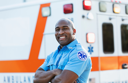 A paramedic standing in front of an ambulance, looking at the camera. He is an African-American man in his 30s.