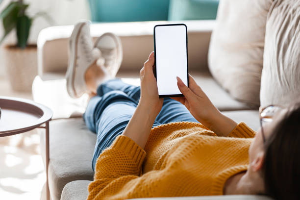 Young woman hold mobile phone in hands, lying on the sofa Young woman hold a mobile phone in hands, lying on the sofa hand holding phone screen stock pictures, royalty-free photos & images