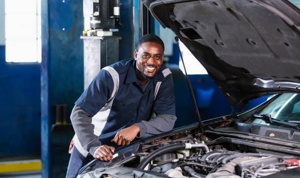 African-American auto mechanic inspecting car engine A mid adult African-American man in his 30s working as a mechanic in an auto repair shop. He is under the hood of a car, inspecting the engine. He is looking at the camera, smiling. mechanic working under car stock pictures, royalty-free photos & images