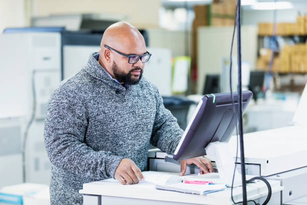 Mixed race man working in printing factory, on computer A mid adult mixed race man in his 30s working in a printing factory. He is using a computer that controls the printing press. digital printing house stock pictures, royalty-free photos & images