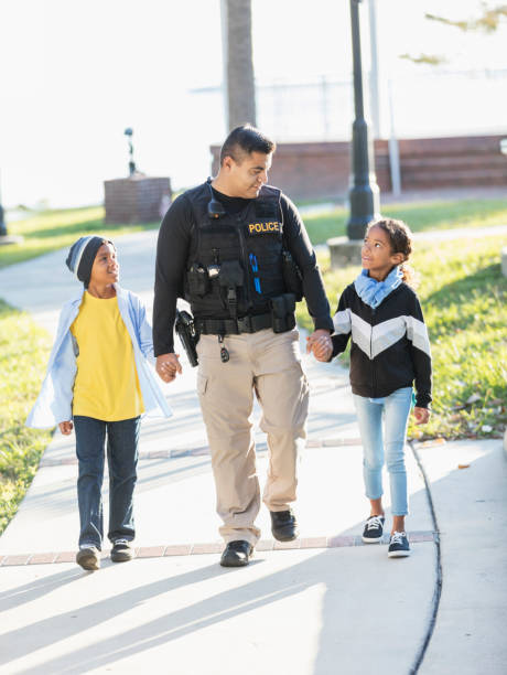 Community policing - an Hispanic police officer is conversing with a girl and her brother, holding hands as they walk. The policeman is a young man in his 20s. The children are 8 and 7 years old, mixed race African-American and Caucasian.