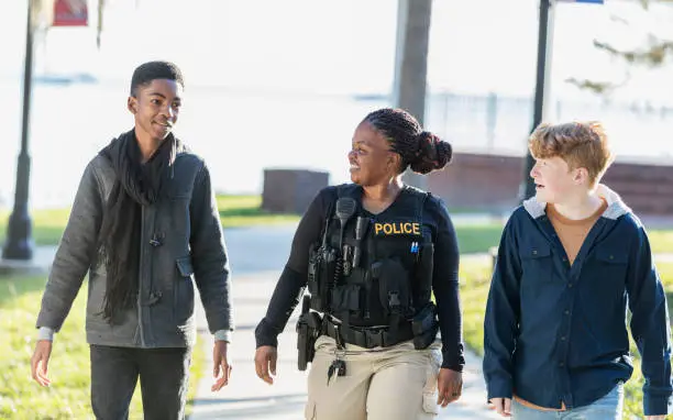 Community policing - a female police officer walking and conversing with two adolescents. The African-American boy is a 14 year old teenager. His friend is 12 years old. The policewoman is a mature African-American woman in her 40s.