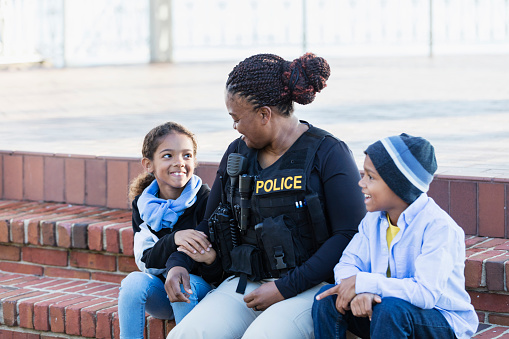 Community policing - a female police officer is conversing with a boy and his sister, sitting side by side on steps outside a building. The policewoman is African-American, in her 40s. The children are 8 and 7 years old, mixed race African-American and Caucasian.