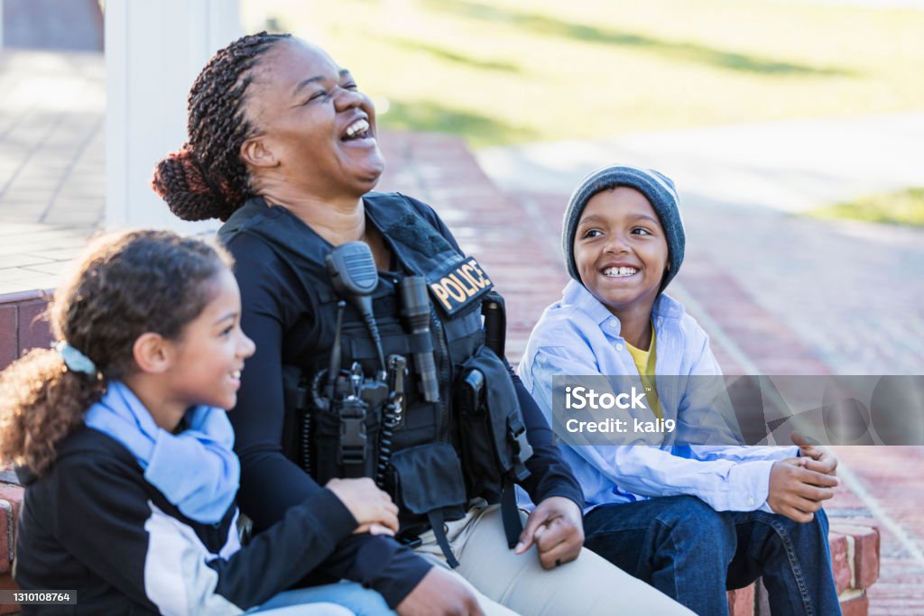 Policewoman in the community, sitting with two children Community policing - a female police officer is conversing with a boy and his sister, sitting side by side on steps outside a building, laughing. The policewoman is African-American, in her 40s. The children are 8 and 7 years old, mixed race African-American and Caucasian. Police Force Stock Photo