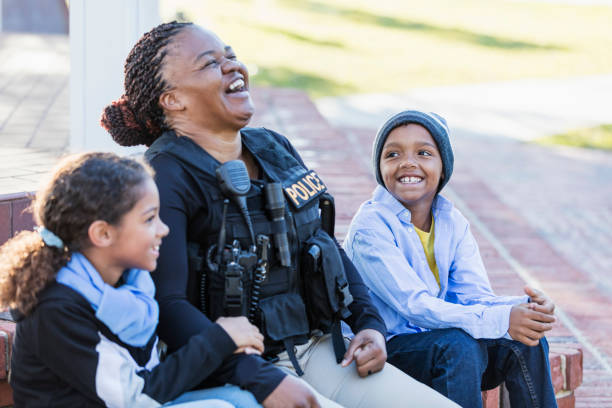 Policewoman in the community, sitting with two children Community policing - a female police officer is conversing with a boy and his sister, sitting side by side on steps outside a building, laughing. The policewoman is African-American, in her 40s. The children are 8 and 7 years old, mixed race African-American and Caucasian. police force smiling officer stock pictures, royalty-free photos & images