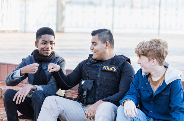 Police officer in community, sitting with two youths Community policing - an Hispanic police officer conversing with two adolescents, and bumping fists. The African-American boy is a 14 year old teenager. His friend is 12 years old. They are sitting side by side on steps outside a building. cheerful kids sitting on steps and gesturing stock pictures, royalty-free photos & images
