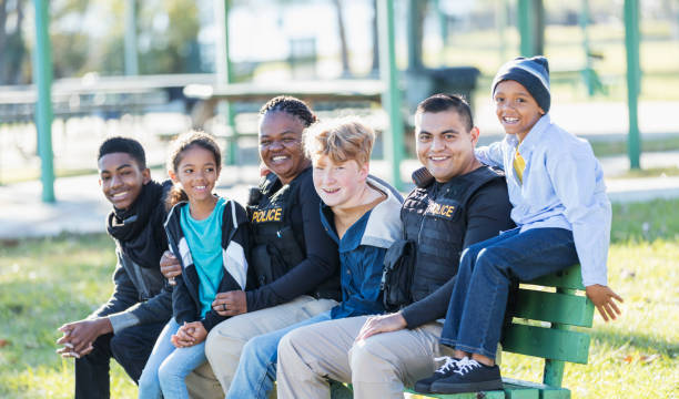 Two police officers sitting with children on park bench Community policing - two police officers hanging out with a group of four multi-ethnic children, sitting side by side on a park bench. The officers are an African-American woman in her 40s and an Hispanic man in his 20s. The children range in age from a 7 year old girl to a 14 year old teenage boy. park bench photos stock pictures, royalty-free photos & images