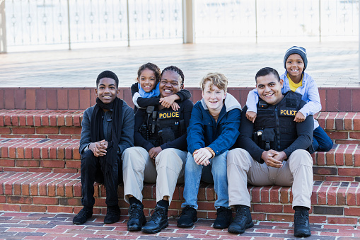 Community policing - two police officers hanging out with a group of four multi-ethnic children, sitting side by side on steps outside a building. The officers are an African-American woman in her 40s and an Hispanic man in his 20s. The children range in age from a 7 year old girl to a 14 year old teenage boy.