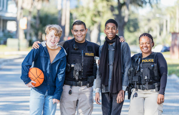 Police officers and two youths with basketball Community policing - two police officers standing with two youths in a public park. One officer, a young Hispanic man, is standing between the two boys, an African-American 14 year old teenager, and a 12 year old boy holding a basketball. The other police officer is an African-American woman. They are smiling at the camera. police force smiling officer stock pictures, royalty-free photos & images