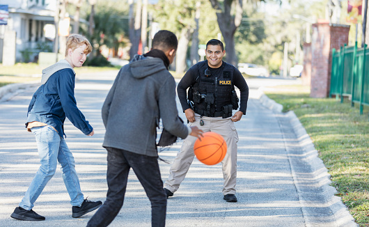 Community policing - a young Hispanic police officer playing basketball with an African-American teenage boy and his friend in a public park.