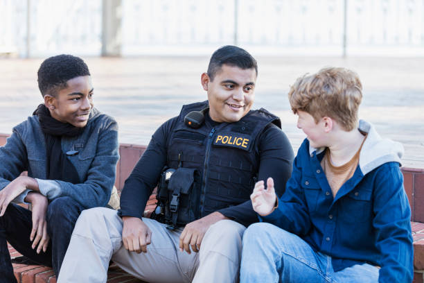 Police officer in community, sitting with two youths Community policing - an Hispanic police officer conversing with two adolescents. The African-American boy is a 14 year old teenager. His friend is 12 years old. They are sitting side by side on steps outside a building. police force smiling officer stock pictures, royalty-free photos & images