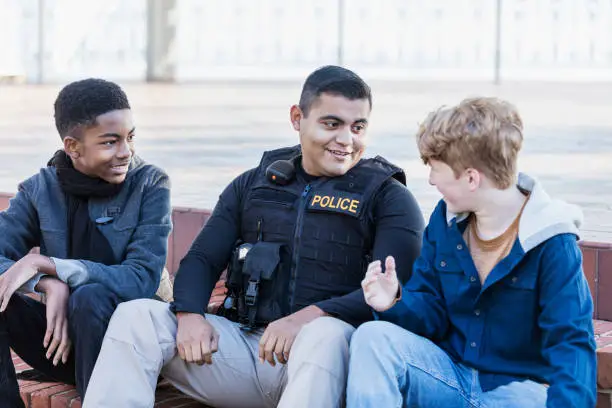 Community policing - an Hispanic police officer conversing with two adolescents. The African-American boy is a 14 year old teenager. His friend is 12 years old. They are sitting side by side on steps outside a building.