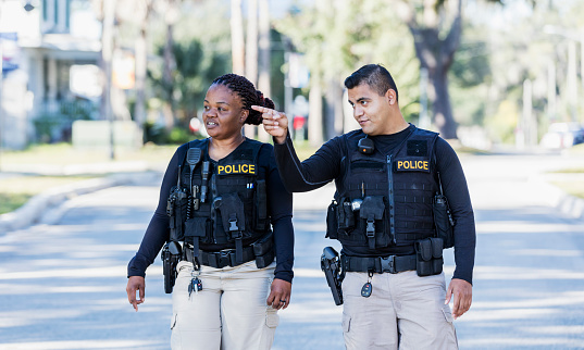 Community policing - two multi-ethnic police officers patrolling a local neighborhood on foot. They are walking side by side. The African-American policewoman is in her 40s. Her partner, a young Hispanic man in his 20s, is pointing at something and they are both looking toward it.