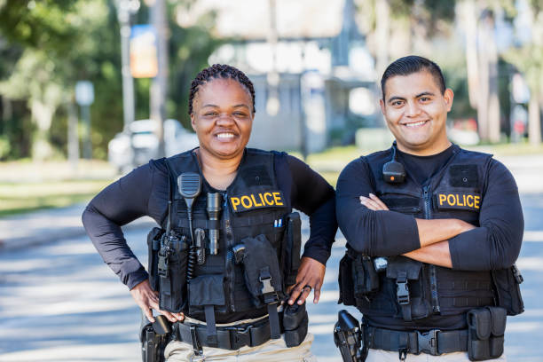 Two police officers on foot patrol Community policing - two multi-ethnic police officers patrolling a local neighborhood on foot. They are standing side by side, smiling at the camera. The African-American policewoman is in her 40s. Her partner is a young Hispanic man in his 20s. police force smiling officer stock pictures, royalty-free photos & images