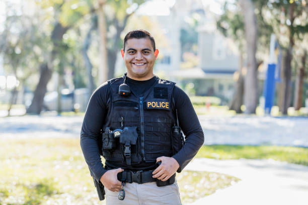 Hispanic police officer on foot patrol Community policing - an Hispanic police officer in his 20s patrols a local neighborhood on foot. He is looking at the camera, smiling. police force smiling officer stock pictures, royalty-free photos & images