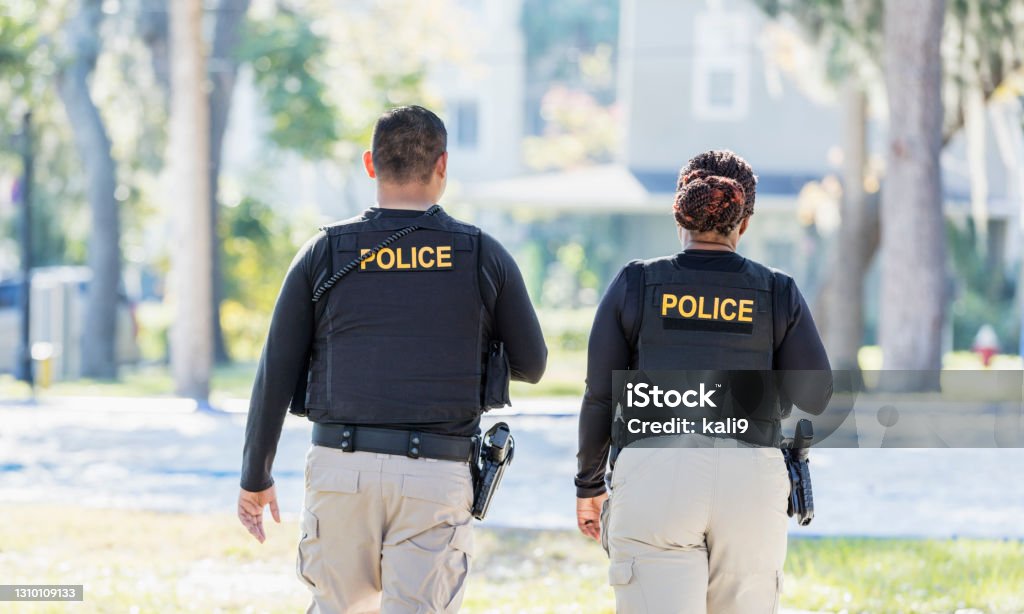 Two police officers walking in community Community policing - rear view of two multi-ethnic police officers patrolling a local neighborhood on foot. They are walking side by side. The African-American policewoman is in her 40s. She and her partner, a young Hispanic man in his 20s. Police Force Stock Photo