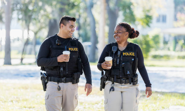 Two police officers walking in community Community policing - two multi-ethnic police officers patrolling a local neighborhood on foot. They are walking side by side, conversing, drinking coffee. The African-American policewoman is in her 40s. She and her partner, a young Hispanic man in his 20s. police force smiling officer stock pictures, royalty-free photos & images