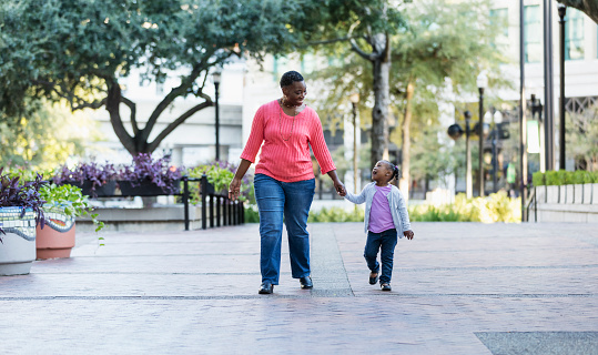 An African-American mother and her little girl walking, holding hands outdoors in a city park. They are looking at each other. Mom is smiling and the little girl is talking with her mouth wide open.