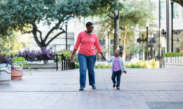 African-American mother and daughter walk in city park An African-American mother and her little girl walking, holding hands outdoors in a city park. They are looking at each other. Mom is smiling and the little girl is talking with her mouth wide open. family in city stock pictures, royalty-free photos & images