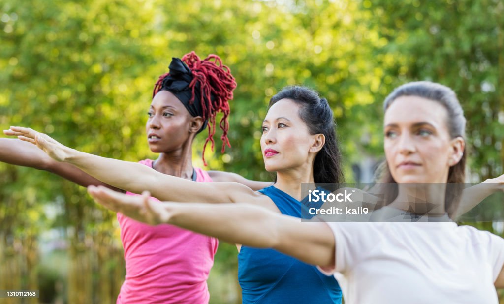 Three multi-ethnic woman in yoga class outdoors A multi-ethnic group of three women in the park taking a yoga class. They are standing side by side, arms outstretched in the warrior position. The focus is on the mature Asian woman in the middle, in her 40s. Her friends are in their 30s. Mature Women Stock Photo