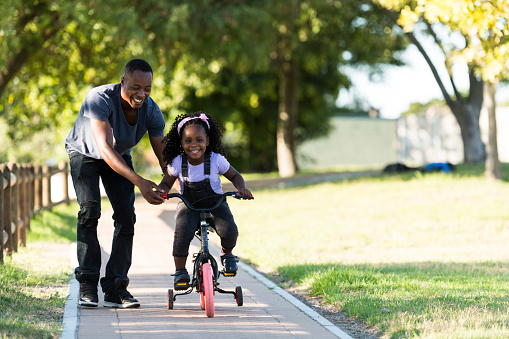 Front view father helps excited little girl riding tricycle