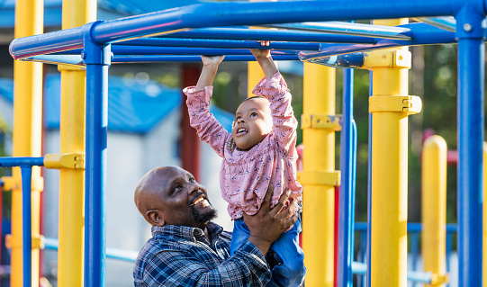 An African-American father playing with his 5 year old daughter at a playground. The little girl has caudal regression syndrome, a rare congenital disorder which affects the development of the lower spine. Dad is helping her play on the monkey bars.