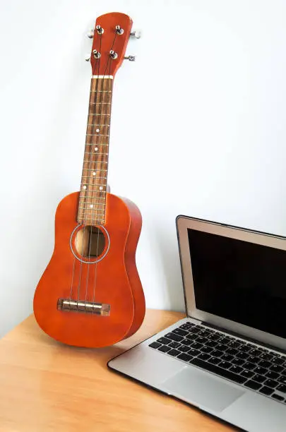 Photo of Hawaiian four-string guitar Ukulele on wooden table and laptop. Concept of learning to play musical instrumen