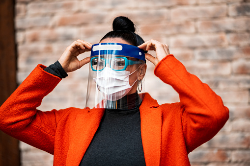 Woman fixing her protective plastic face shield over the face mask and eyeglasses that she is wearing as a supreme safety measure during the pandemic.
