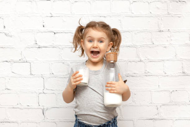 surprised cute child girl with expression face and mustache of milk on the lips over white brick wall holding glass with milk and bottle of milk.banner, copy space for text, mock up - milk mustache imagens e fotografias de stock