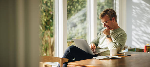 Young man using laptop and smiling Young man using laptop and smiling at home. Man sitting by table working on laptop computer. working from home stock pictures, royalty-free photos & images