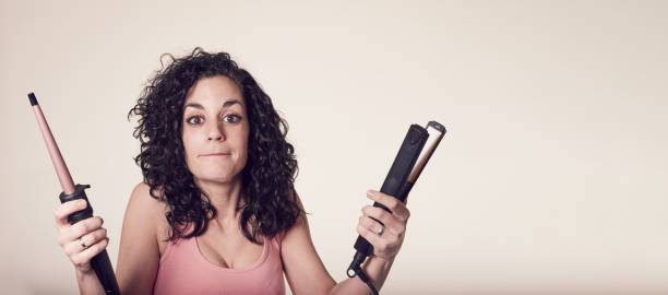 Young smiling curly-haired woman can't decide between using her curling iron or her hair straightener. doubt gesture. care and beauty concept. Young smiling curly-haired woman can't decide between using her curling iron or her hair straightener. doubt gesture. care and beauty concept. curling tongs photos stock pictures, royalty-free photos & images