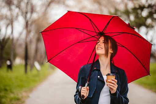 Portrait of smiling young woman enjoying coffee and walking in public park with red umbrella.