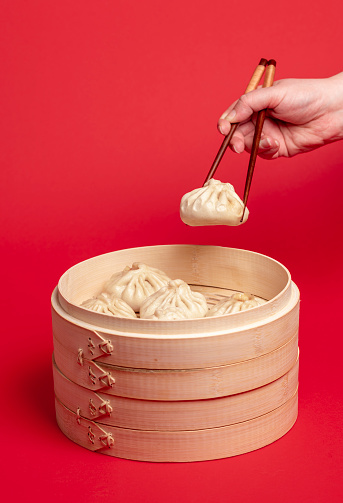 Woman taking a dumpling from the bamboo steamer with the chopsticks. Freshly steamed baozi dumplings in a wooden steamer isolated on a red colored background.