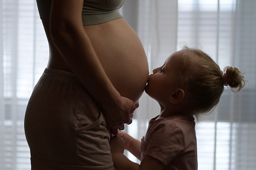 Little girl kissing her young mom's pregnant belly on a background of a window. Image of pregnancy and expectation of a child
