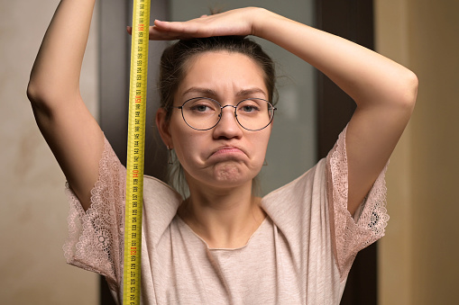 A young woman shows sadness at her height by holding a measuring tape next to her. The growth of a short woman and negative emotion