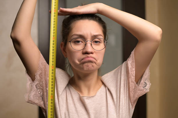 A young woman shows sadness at her height by holding a measuring tape A young woman shows sadness at her height by holding a measuring tape next to her. The growth of a short woman and negative emotion woman taller stock pictures, royalty-free photos & images