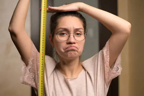 A young woman shows sadness at her height by holding a measuring tape next to her. The growth of a short woman and negative emotion