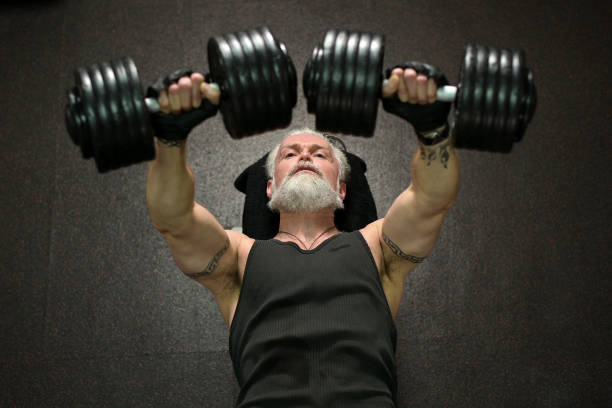 An overhead view of an athletic gray-haired man with a beard and tattoos, training his chest muscles An overhead view of an athletic gray-haired man with a beard and tattoos, pressing dumbbells on a bench, training his chest muscles in the gym senior bodybuilders stock pictures, royalty-free photos & images