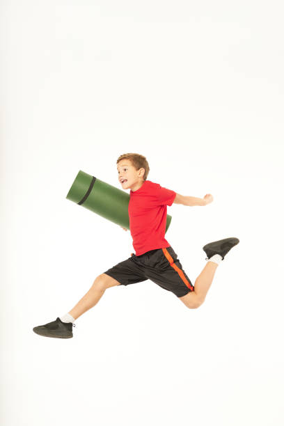 Cute male child in sportswear holding rolled exercise mat and jumping in the air. Isolated on white background
