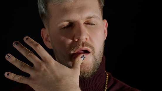Close-up shot of confident man in stylish clothing sensually touching lips with finger on black background. Facial expression of trendy bearded guy with gray hair. People lifestyle. Modern fashion