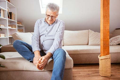 Senior man suffering with foot cramp on sofa in living room at home.